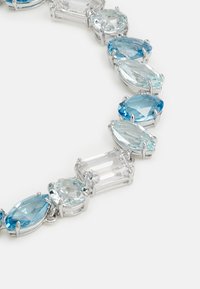 Piedras preciosas azules y transparentes en varias formas, montadas en metal de tono plateado. El diseño alterna entre piedras ovaladas y rectangulares.