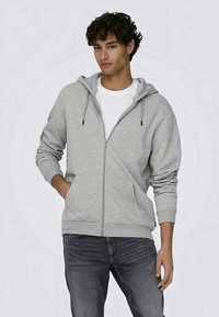 Felpa grigia con zip e cappuccio regolabile, due tasche frontali, polsini e fondo a coste, indossata sopra una t-shirt bianca, abbinata a jeans scuri.