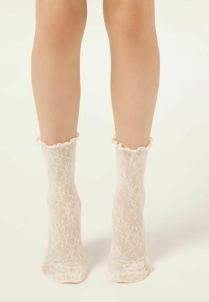 Beine, die durchsichtige weiße Spitzen-Knöchelsocken mit Blumenmuster und Rüschenrändern tragen, stehend auf hellem Hintergrund.