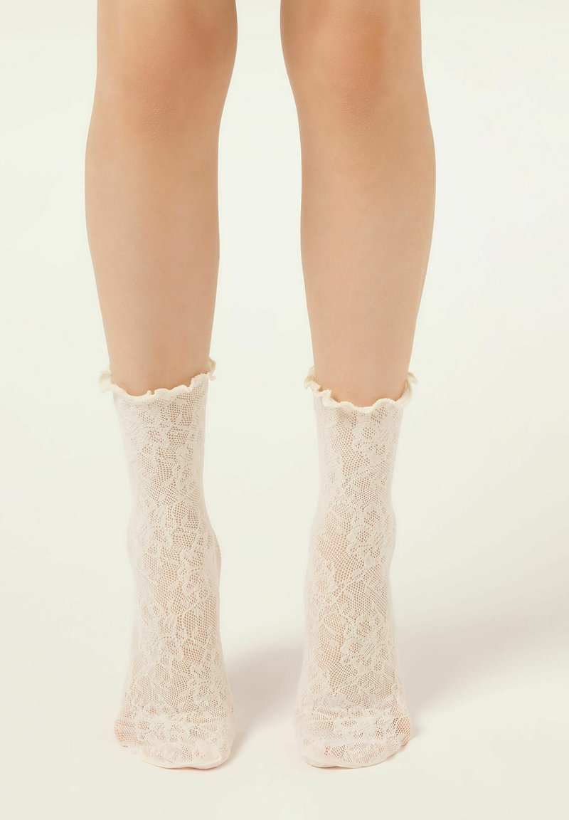 Jambes portant des chaussettes de cheville en dentelle blanche transparente avec motif floral et bords froncés, debout sur un fond clair.