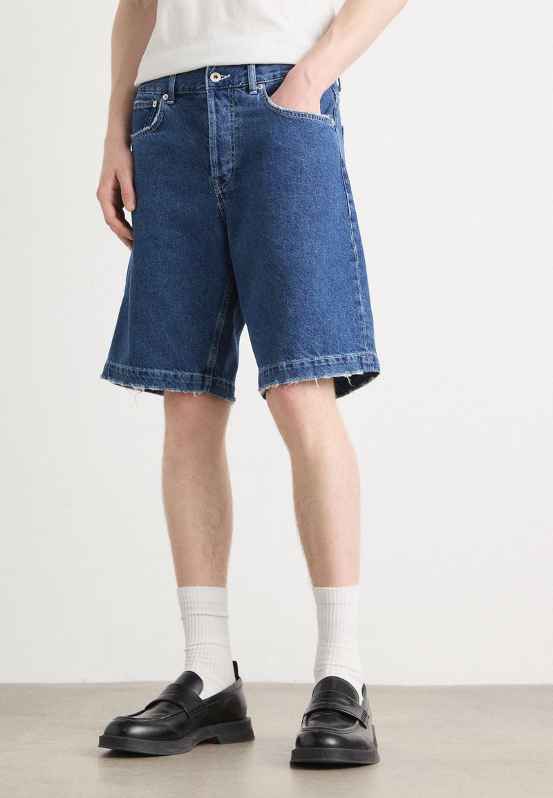 Shorts en jean bleu foncé avec une coupe décontractée, des bords effilochés et cinq poches. Assortis avec des mocassins noirs et des chaussettes blanches montantes.
