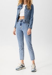 Donna che indossa una giacca di jeans cropped, una maglietta bianca cropped, jeans azzurri a vita alta e sneakers blu e bianche, in piedi su uno sfondo bianco.