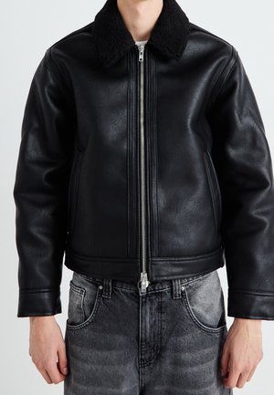 Mężczyzna w czarnej skórzanej kurtce z czarnym kołnierzem z owczej skóry, z zamkiem z przodu i wyblakłych czarnych dżinsach, stojący na jednolitym tle.