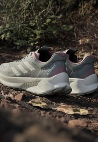 Scarpe da outdoor Adidas Terrex grigie e rosa con una parte superiore testurizzata, tre strisce bianche e una suola bianca spessa. Presenta un tallone rinforzato.