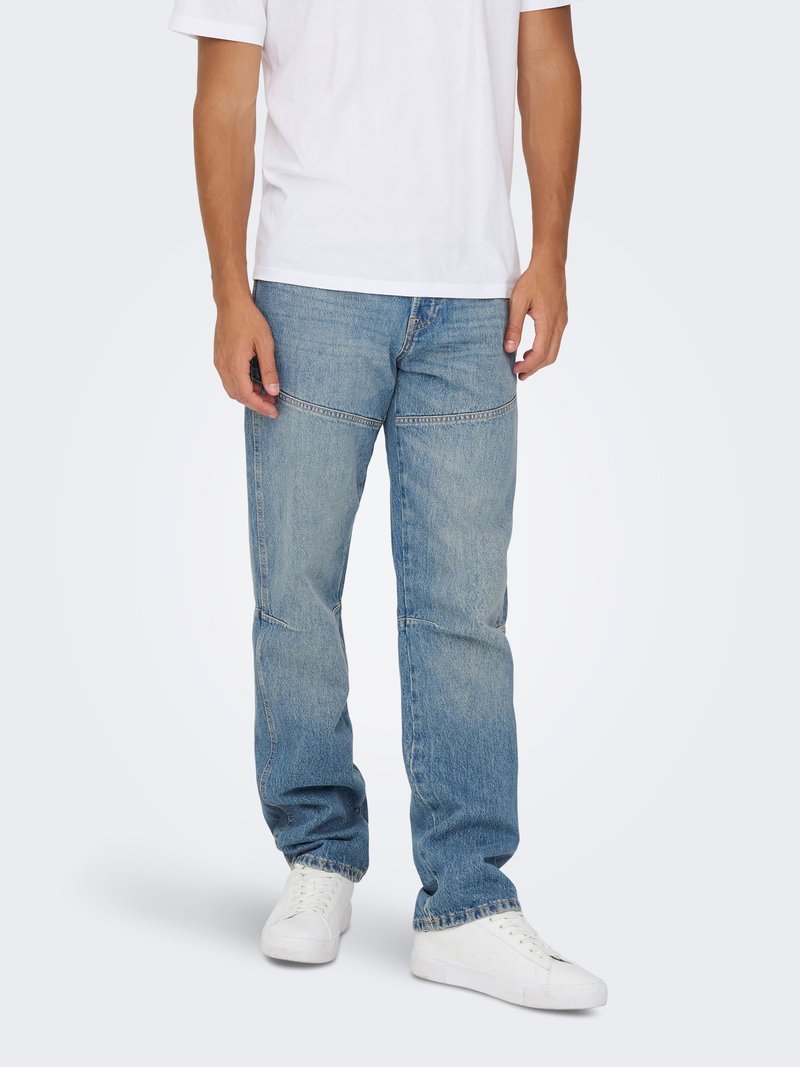 Mann trägt locker sitzende blaue Jeans mit horizontalen Nahtdetails und weißen Sneakers, steht vor einem einfarbigen Hintergrund.