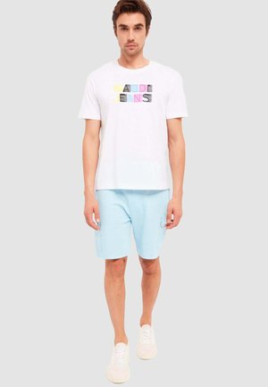 Maglietta bianca con grafica colorata "CALIJEANS", abbinata a pantaloni corti azzurri chiari con tasche laterali e sneaker bianche. Design semplice.