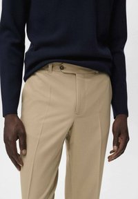 Pantalones tailored beige con una textura suave, que cuentan con un bolsillo frontal, cierre de botón y pliegue vertical. Llevados con un suéter azul marino.