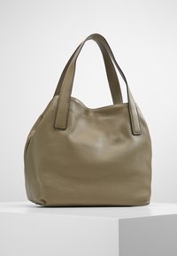 Miękki oliwkowozielony skórzany torebka na ramię z dwoma szerokimi paskami, wyeksponowana na białym podwyższeniu na tle jasnoszarym.