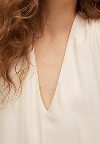 Lichtbeige blouse met een diepe V-hals. De stof heeft een soepele textuur, valt zacht met subtiele contouren rond de kraag.