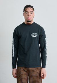 Black long-sleeve shirt with white "OBEY" logo and text on sleeves. Worn with brown checkered pants. Smooth fabric, round neck.