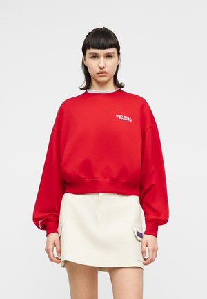 Młoda kobieta z krótkimi ciemnymi włosami nosi czerwoną bluzę z napisem „Red Bull Racing” i jasnobeżową spódnicę na gładkim szarym tle.