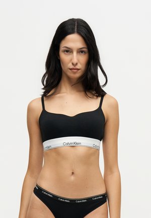 Kvinde med sort hår iført en sort bh og sort undertøj med hvide Calvin Klein-logo bånd på en ensfarvet lys baggrund.