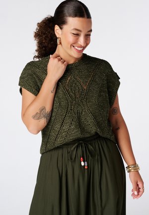 Vrouw glimlachend in een donkergroene gebreide top en bijpassende losse broek met kralenkoord, toont tatoeages op beide armen en draagt gouden sieraden.