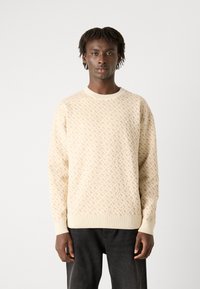 Pulover crem cu un model texturat de tricot și manșete și tiv de un bej deschis în contrast. Asortat cu blugi negri.
