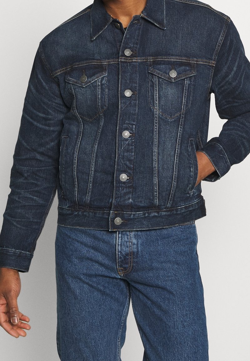 Dark blue denim jacket, featuring button-up front, two chest pockets, and side pockets. Fitted design with contrasting stitch details.