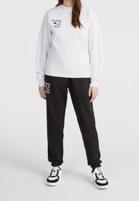Mujer vestida con un sudadera blanca y pantalones de chándal negros con el texto "Women of the Wave", combinados con zapatillas en blanco y negro, de pie sobre un suelo blanco.