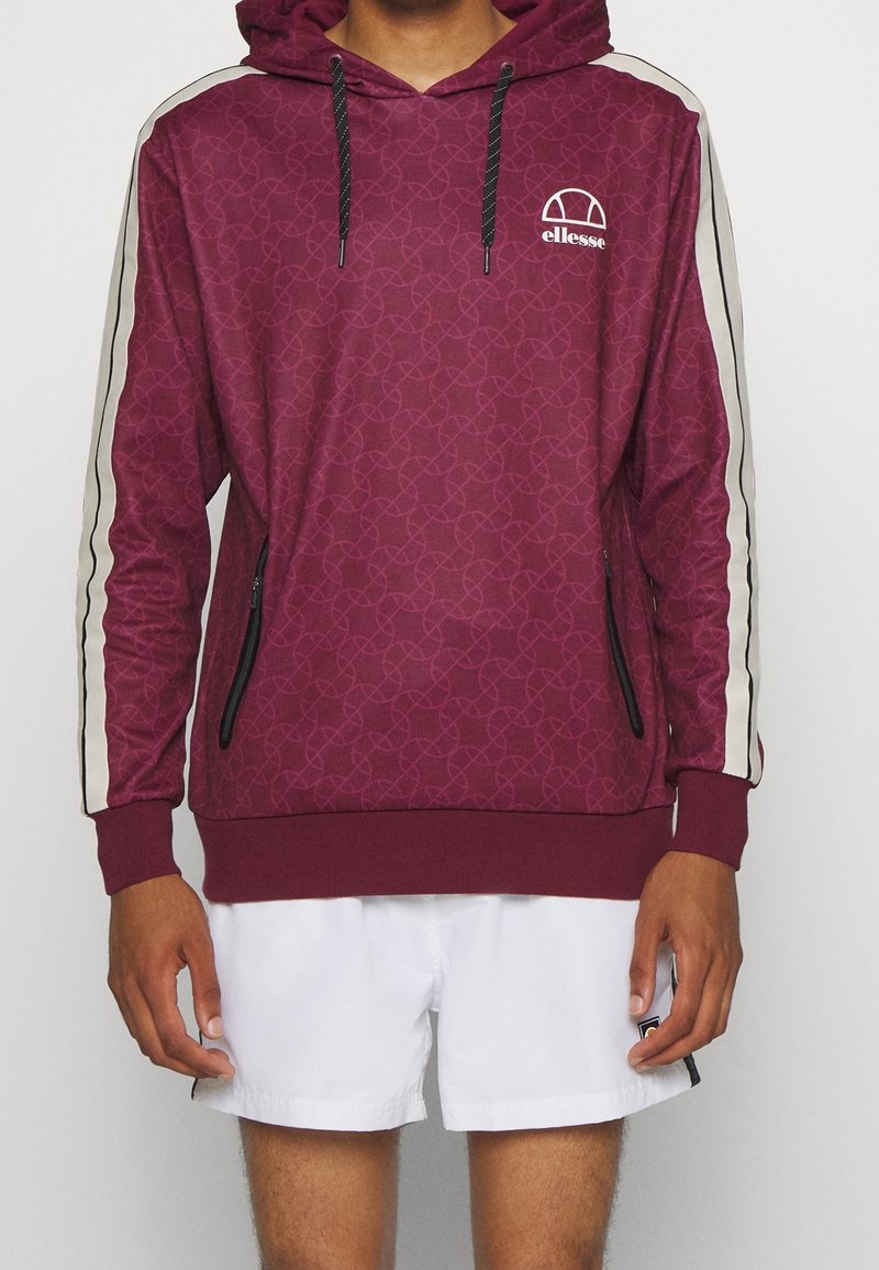 Homme portant un sweat à capuche bordeaux avec des bandes blanches sur les côtés et des cordons noirs, associé à un short blanc, debout devant un fond uni.