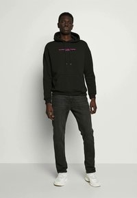Uomo che indossa un hoodie nero con testo rosa, jeans scuri e sneaker bianche, in piedi contro uno sfondo chiaro uniforme.