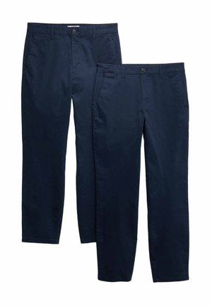 Man met een marineblauwe jas, crêmekleurige trui, donkerblauwe broek, bruine riem en bruine schoenen, staand met handen in de zakken, kijkt opzij.