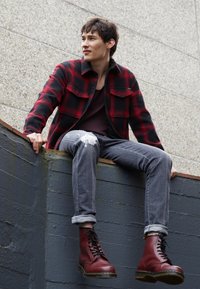 Jeune homme portant une veste à carreaux rouge et noire, un jean gris déchiré et des bottes rouges à lacets, assis sur un rebord devant un mur texturé.