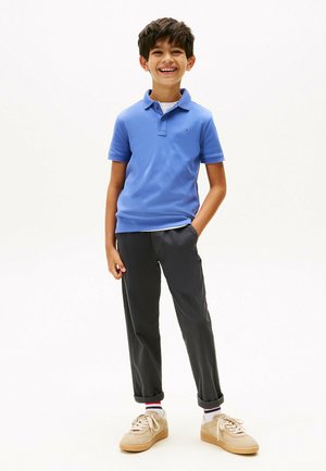 Polo blu con colletto e maniche corte, abbinato a pantaloni scuri e sneakers beige. Presenta un piccolo logo sulla maglietta e le maniche arrotolate.