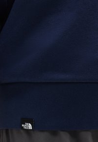 Ναυτικό μπλε ύφασμα με ραμμένη μαύρη ετικέτα όπου φαίνεται το λευκό λογότυπο «The North Face» κοντά στην κάτω άκρη.