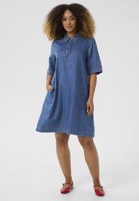 Denim jurk in blauw, met korte mouwen, een knoopsluiting en zijzakken. De zoom heeft een licht rafelige rand. Gemaakt met rode flats.