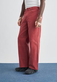 Κόκκινο τζιν με φαρδύ παντελόνι, φτιαγμένο από denim, με κουμπί και φερμουάρ, ευθείες ραφές και φθαρμένη υφή. Συνδυασμένο με μαύρα αθλητικά παπούτσια.