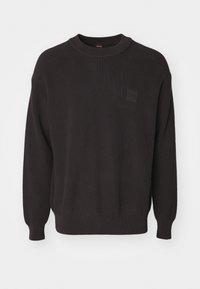 Black knitted sweater with ribbed texture, round crew neckline, and a small square logo patch on the left chest. Long sleeves.