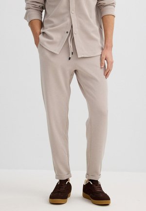 Pantalones joggers beige con textura de canalé, cintura ajustable con cordón, corte slim, dobladillos con puños, combinados con zapatillas de color marrón oscuro.