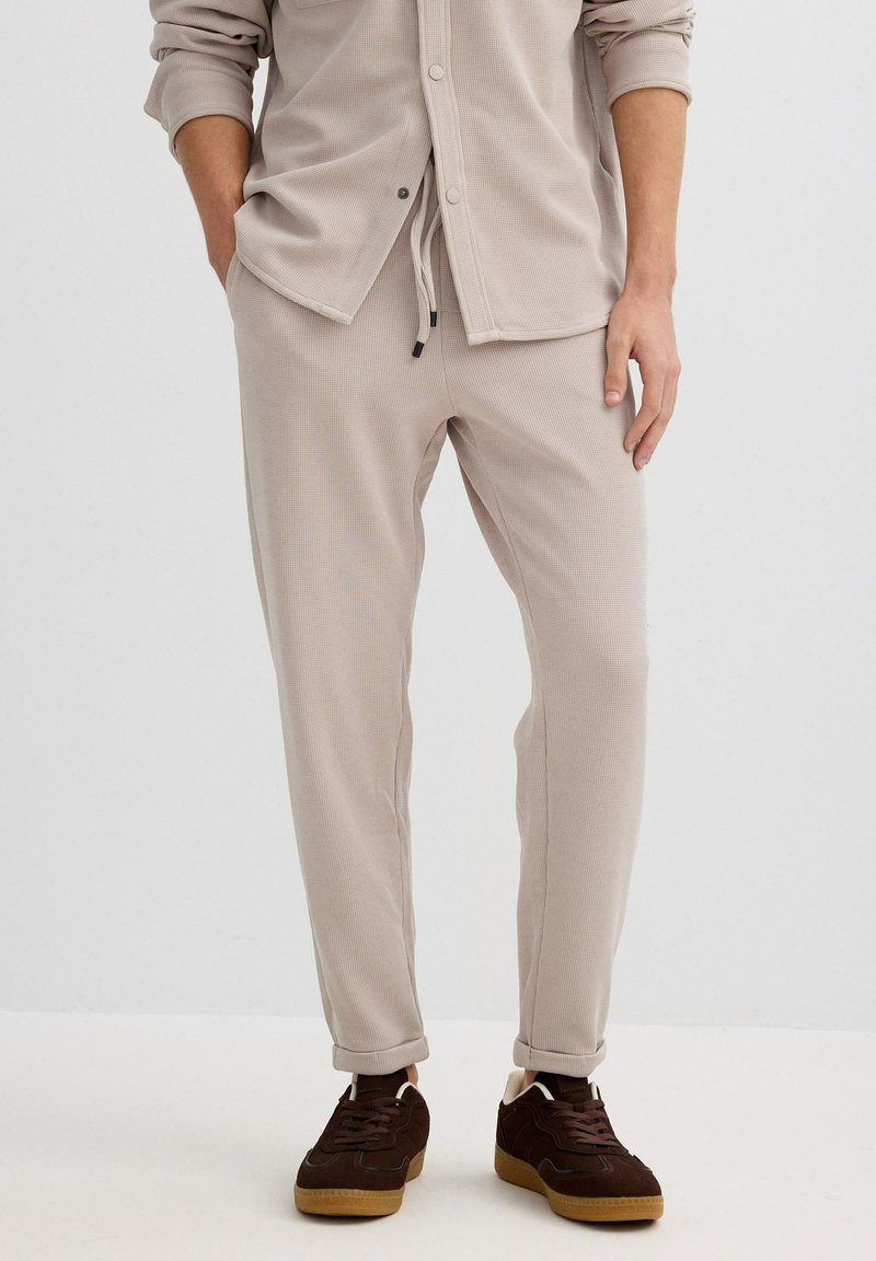 Pantalones joggers beige con textura de canalé, cintura ajustable con cordón, corte slim, dobladillos con puños, combinados con zapatillas de color marrón oscuro.