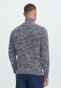 Hombre vestido con un suéter de punto jaspeado en azul marino y blanco con cuello alto, visto desde atrás contra un fondo claro y liso.