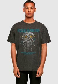 Tricou oversized gri cu o textură estompată, având un grafic cu un schelet și textul albastru "Iron Maiden". Partea din față include detaliul "World Slavery Tour 1984".