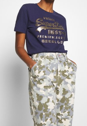 Personne portant un t-shirt bleu marine avec un texte doré et un pantalon camouflage bleu clair, gris et vert olive, les mains dans les poches.