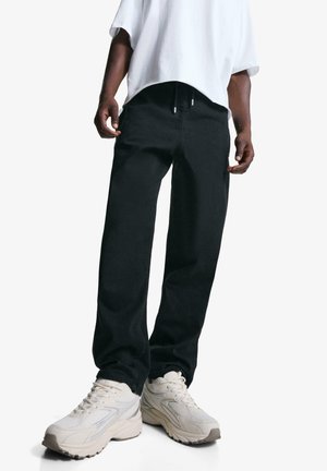 Black denim pants with a relaxed fit and drawstring waist, paired with beige athletic shoes. Smooth texture, simple design, no visible patterns.