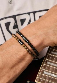 Due braccialetti di perle indossati su un polso. Uno presenta perle nere con un accento lavorato, l'altro ha perle marmorizzate oro e marrone.