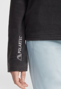 Zwarte fleece pullover met het Polartec-logo op de mouw, voorzien van een zachte textuur en een ontspannen pasvorm, gecombineerd met lichtblauwe broek.