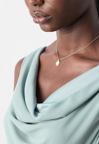 Gros plan sur le cou et les épaules d’une femme portant un haut drapé bleu clair et un délicat collier en chaîne dorée avec un petit pendentif rond.