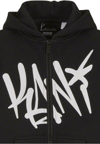 Svart hettejakke med glidelås som har en fremtredende hvit graffiti-inspirert logo på forsiden, med snøring i hetten og myk stofftekstur.