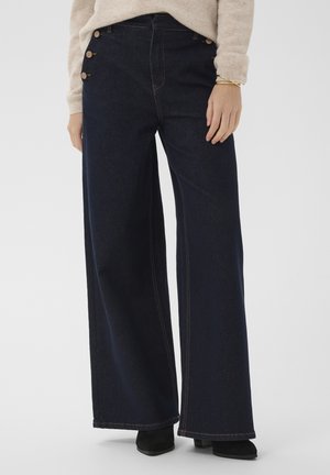 Femme portant un jean large foncé avec des boutons sur le côté, un pull beige, des chaussures noires et un bracelet en or à un poignet.