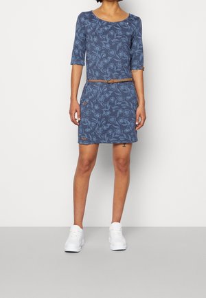 Vrouw die een blauwe bloemrijke jurk tot aan de knieën draagt met driekwartmouwen, een bruine gevlochten riem en witte sneakers, staand tegen een witte muur.