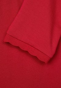 Tessuto di maglione rosso con una texture liscia; presenta un orlo scallopato al polsino e dettagli a coste. Colore morbido e uniforme in tutto.