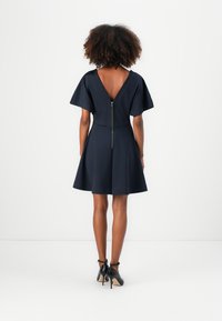 Vestido azul marino con espalda en V, mangas cortas, corsé ajustado y falda amplia. Cuenta con un cierre de cremallera en la parte posterior. Llevado con tacones negros.
