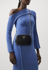 Borsa a tracolla nera trapuntata con dettagli in oro. Caratterizzata da una tracolla a catena e un accento del logo. Indossata con un abito blu a spalle scoperte con coste.