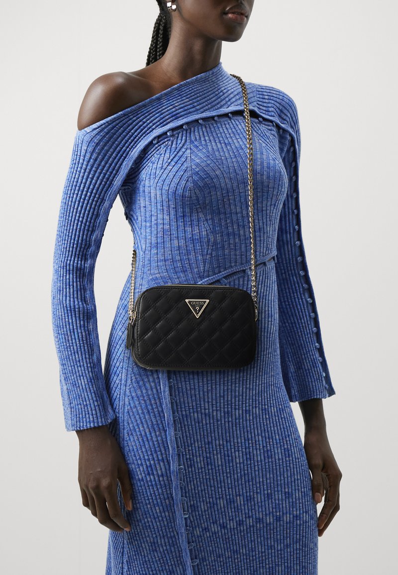 Borsa a tracolla nera trapuntata con dettagli in oro. Caratterizzata da una tracolla a catena e un accento del logo. Indossata con un abito blu a spalle scoperte con coste.