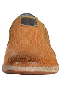 Chaussure brune à enfiler avec une tige texturée et un motif, accents verts contrastants et semelle texturée. Présente un patch logo noir sur le côté.