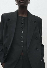Traje gris oscuro a medida con un blazer de doble botón, solapas anchas y una camisera ajustada con botones grandes y brillantes de color negro.