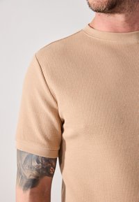 Beige textured short-sleeve shirt with a ribbed design and a round neckline, featuring a visible tattoo on the arm.