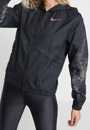 Eine Frau trägt eine schwarze Nike-Jacke mit Reißverschluss und lila Logo sowie gepunktetem Design an den Ärmeln, kombiniert mit dunklen Leggings.