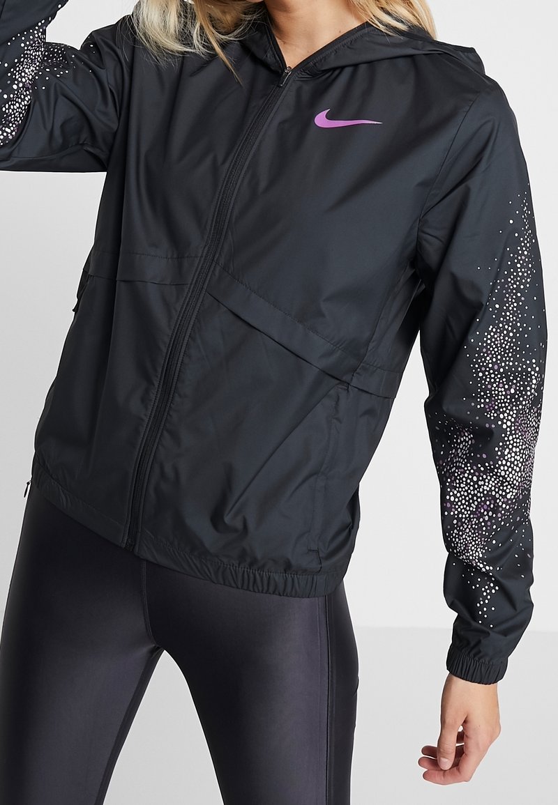 Mujer vestida con una chaqueta con cremallera negra de Nike con un logo morado y un diseño de puntos en las mangas, combinada con leggings oscuros.